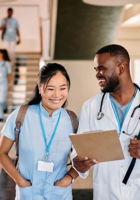 Two doctors in the foreground, a woman of Asian descent on the left wearing scrubs and a name badge turned backward, and a Black man on the right wearing a white lab coat and a stethoscope. Both are smiling, with the man looking at the woman while she looks at the clipboard he is holding.